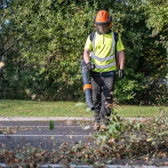 A person wearing safety gear and a high-visibility vest uses a leaf blower to clear leaves and debris from a roadside, with trees and grass in the background.